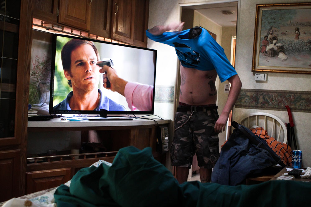 Ryan struggles to get his shirt on by himself while his father watches Dexter on the pullout couch in the camper.